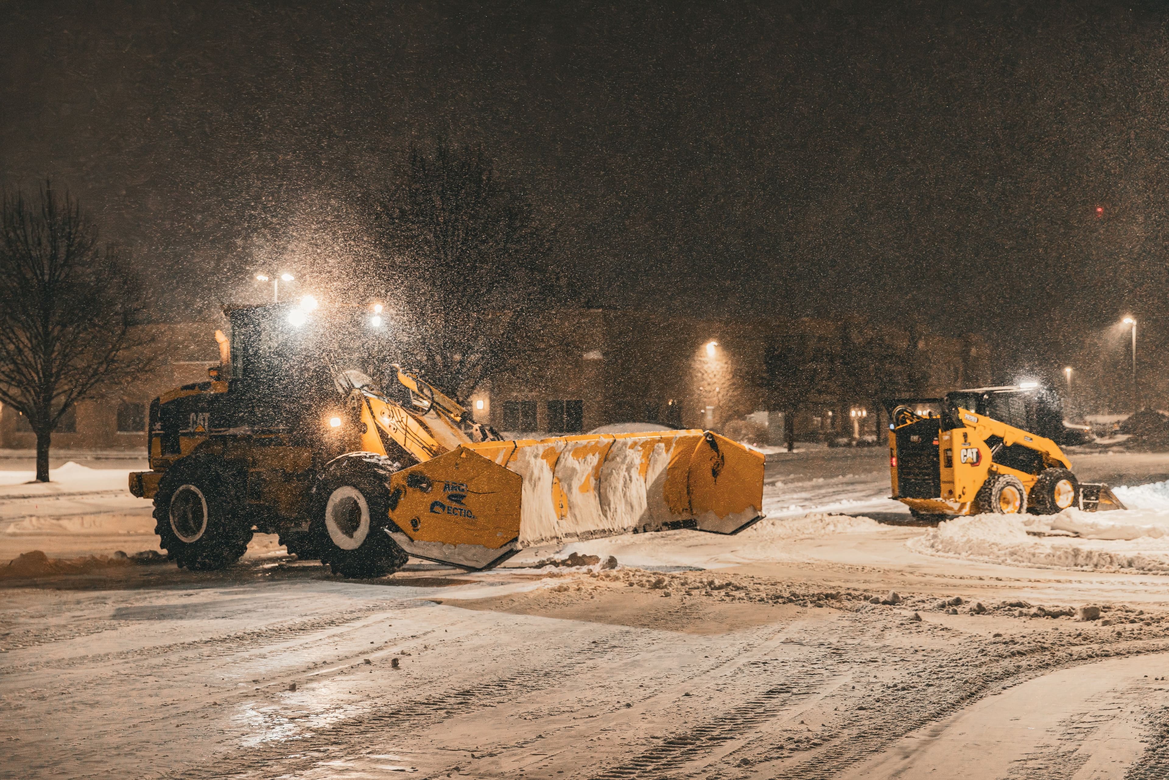 Heavy machinery clearing snow in a parking lot during a nighttime snowstorm.