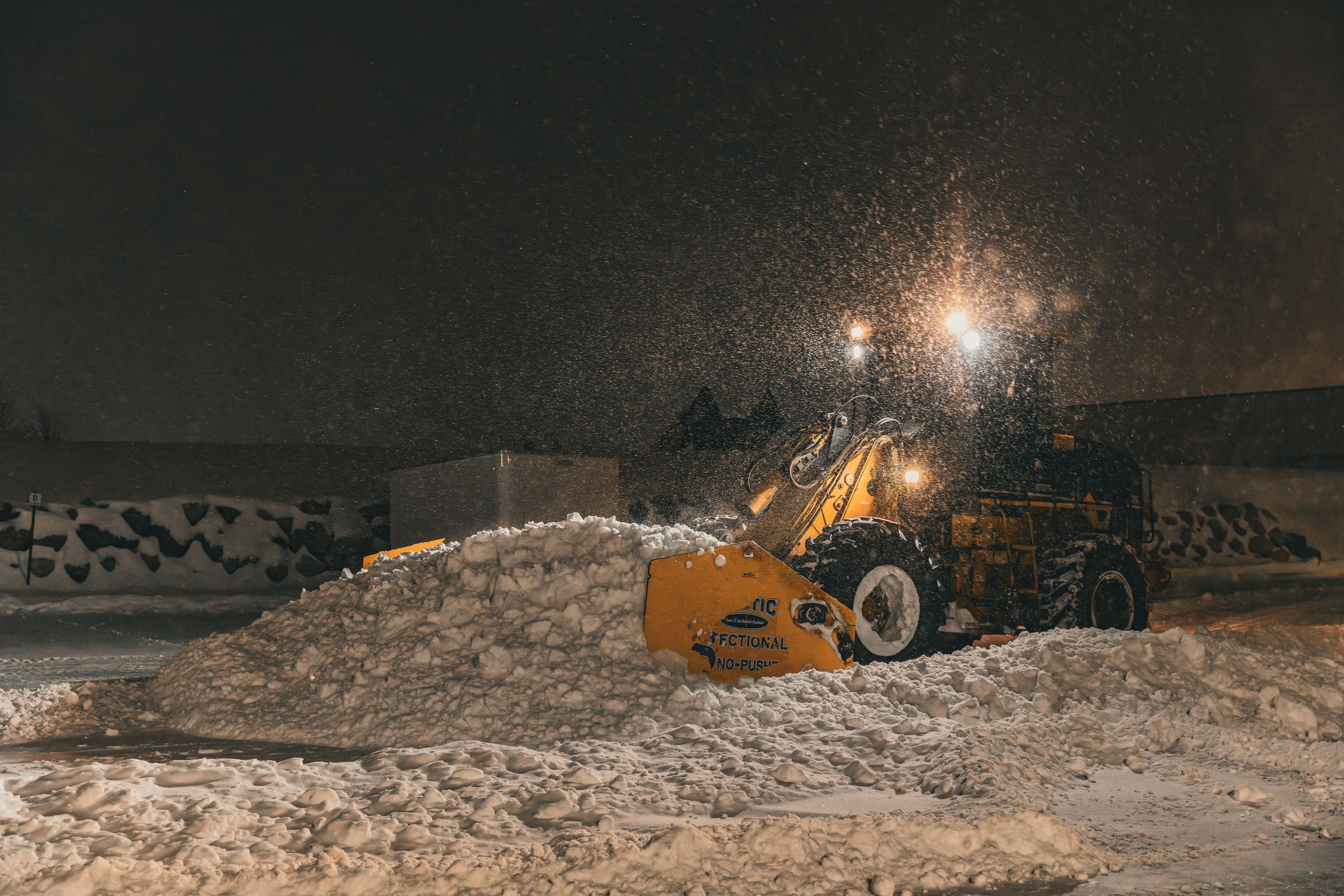Snow plow clearing heavy snow at night with bright headlights illuminating the scene.