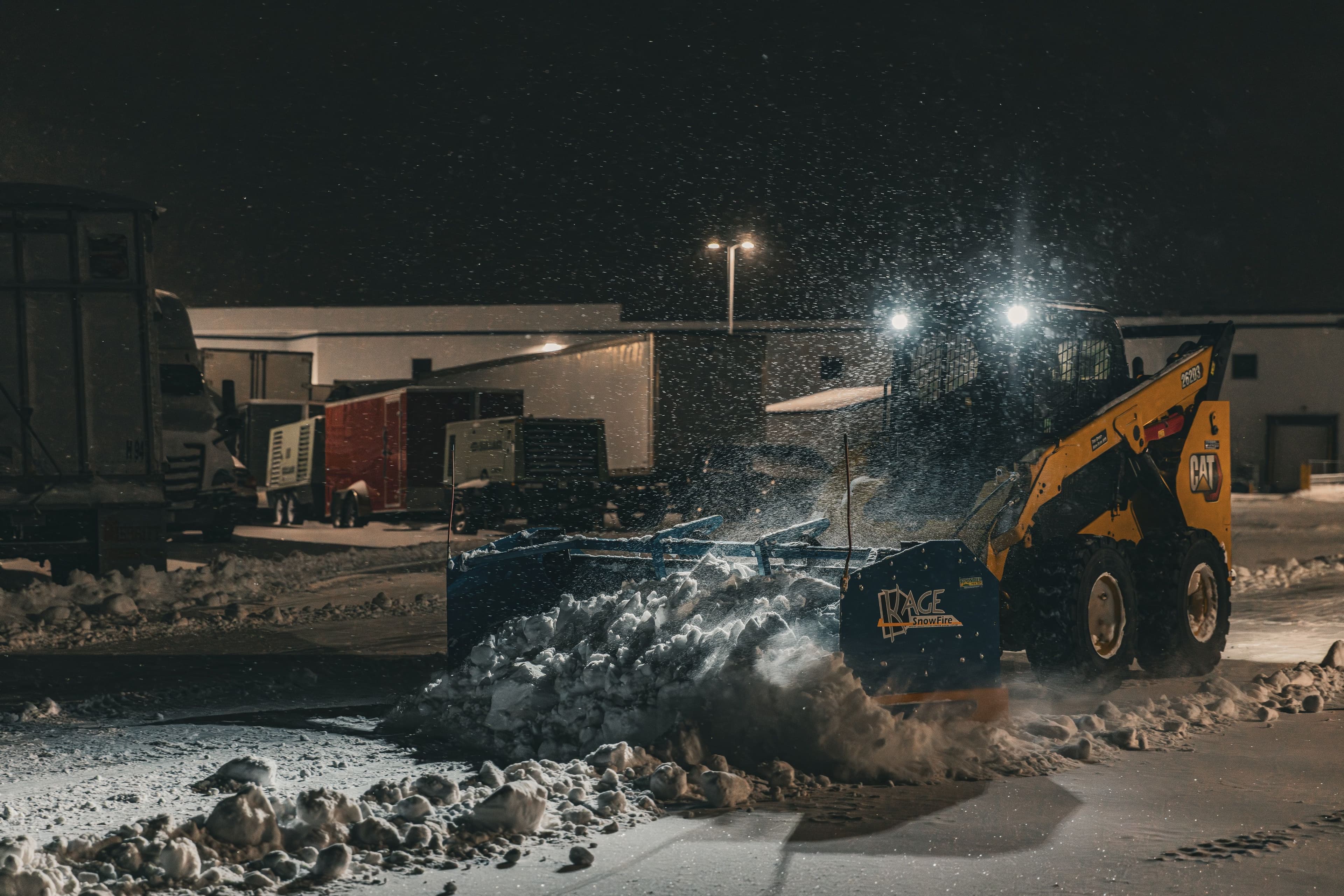 Snow plow clearing snow at night in a commercial lot, illuminated by headlights.