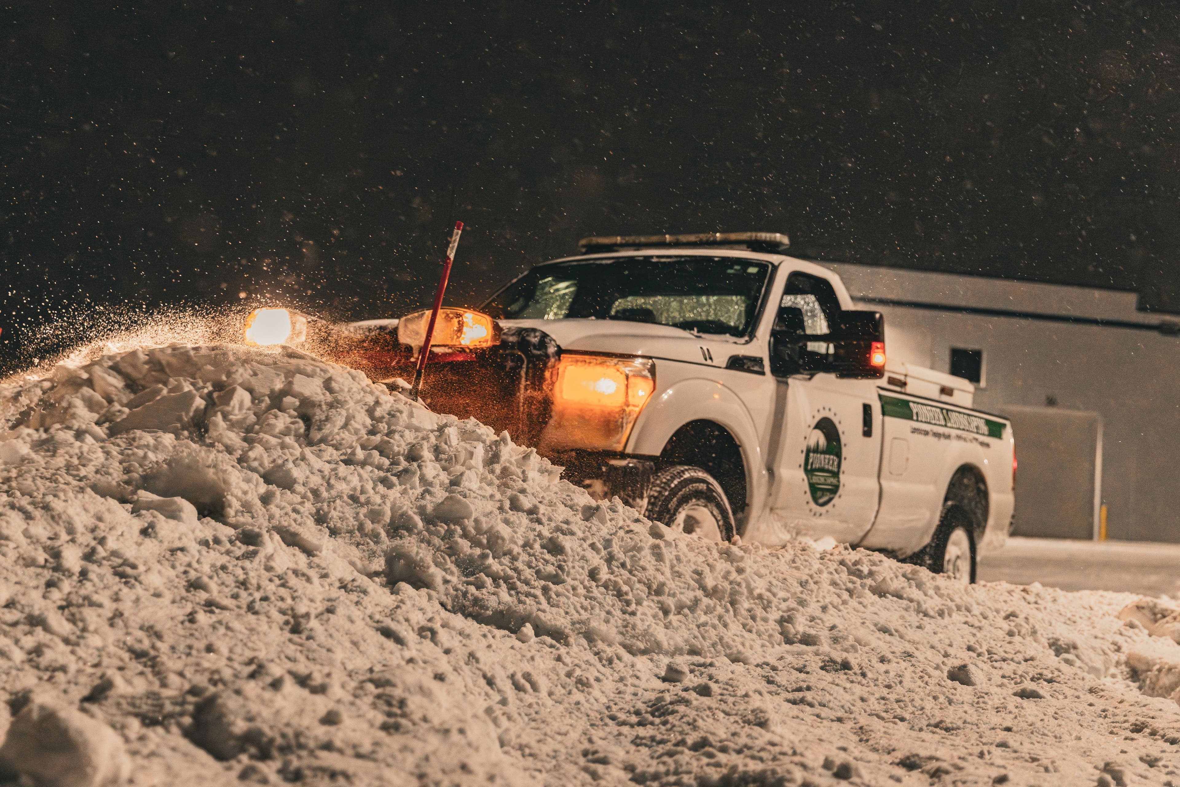 Snow plow truck clearing heavy snowfall at night, with bright headlights illuminating the scene.