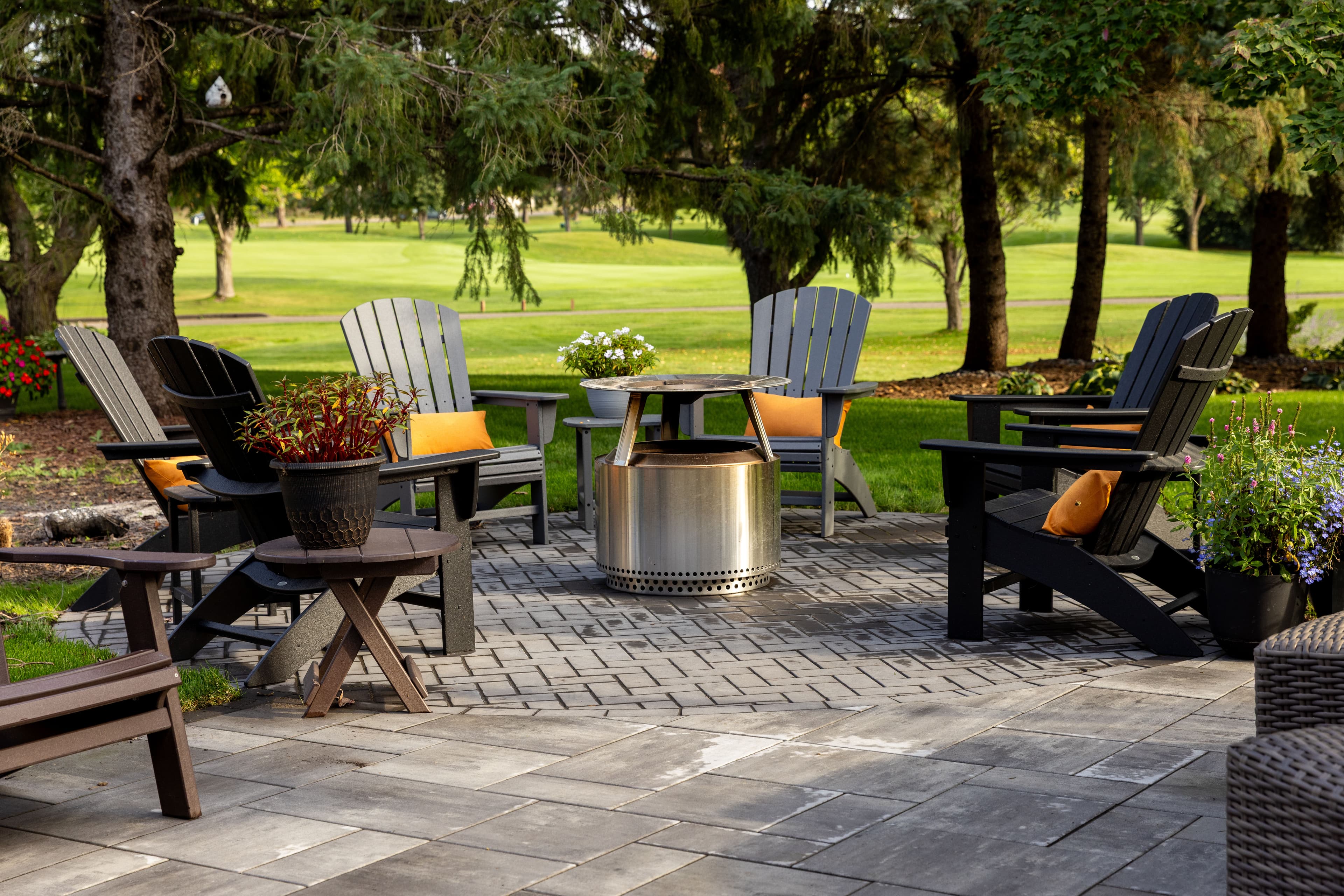 Outdoor seating area with black Adirondack chairs, fire pit, and lush greenery. Ideal for relaxation.