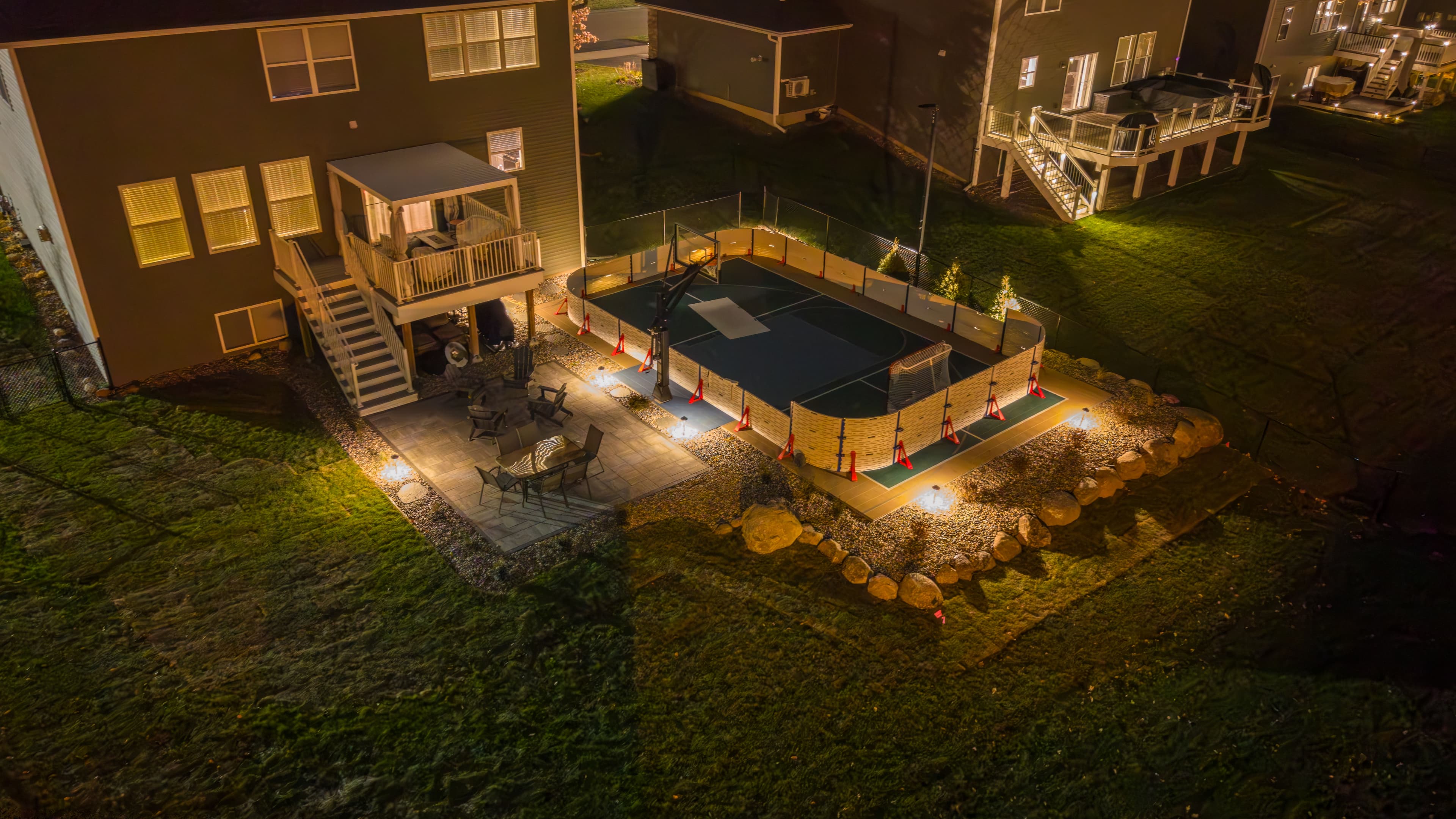 Backyard sports court illuminated at night, surrounded by landscaping and outdoor seating.