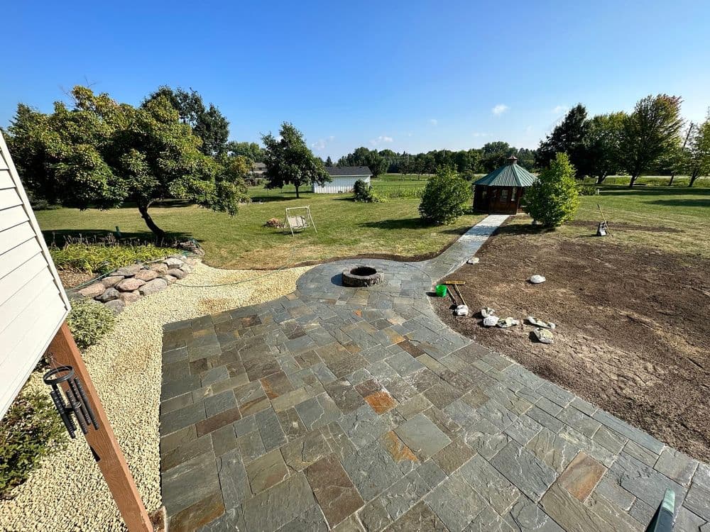 View of a landscaped backyard with a stone patio, fire pit, and gazebo near a lake.