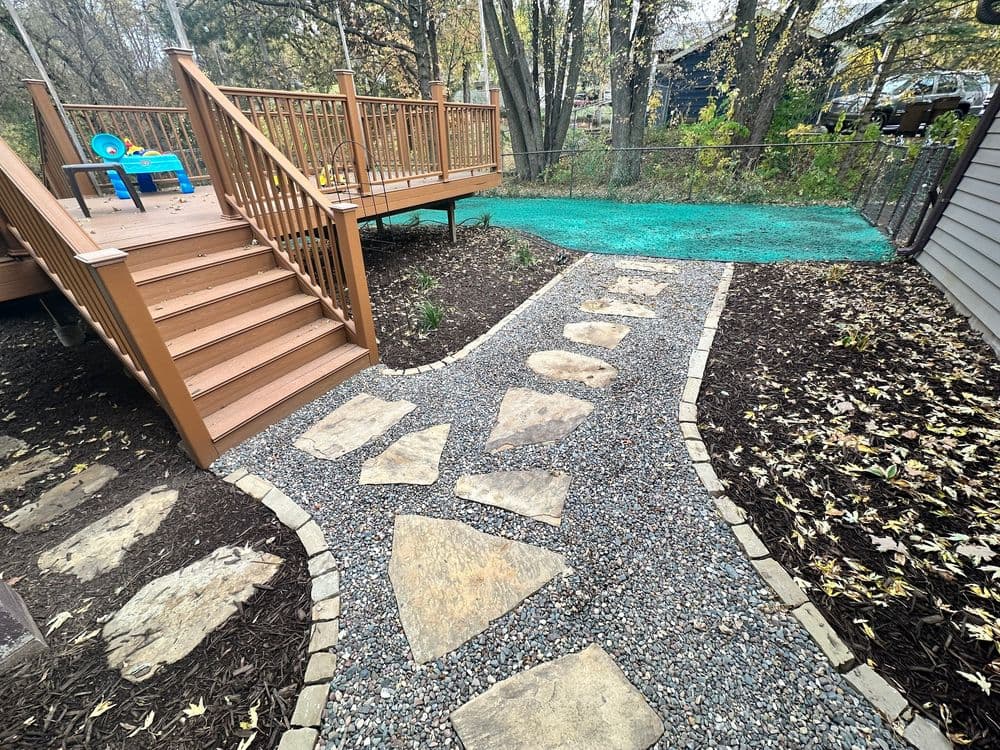 Backyard landscape featuring a stone pathway, wooden deck, and surrounded by greenery.