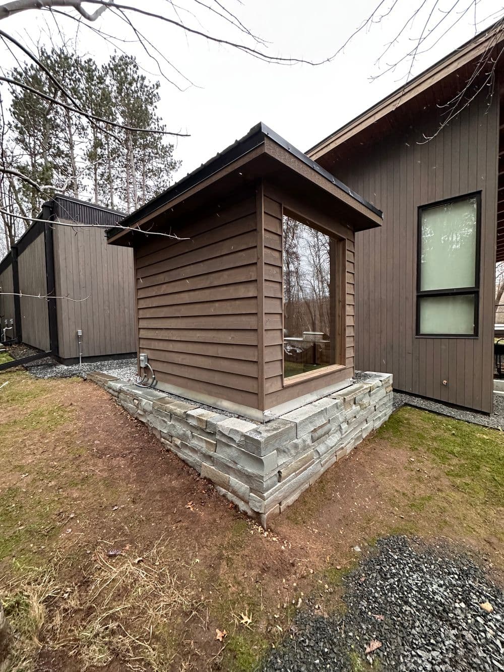 Modern wooden shed with stone base, surrounded by trees and contemporary buildings.