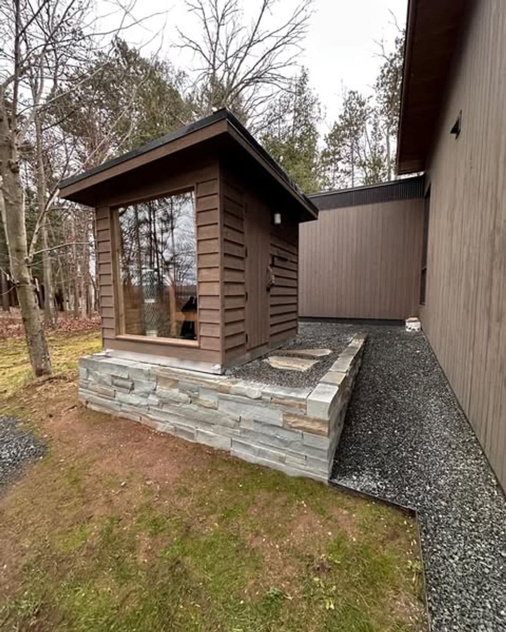 Modern wooden structure with stone foundation beside a contemporary house in a forested area.