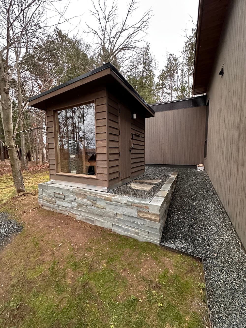 Wooden shed with stone foundation beside modern house in landscaped yard.
