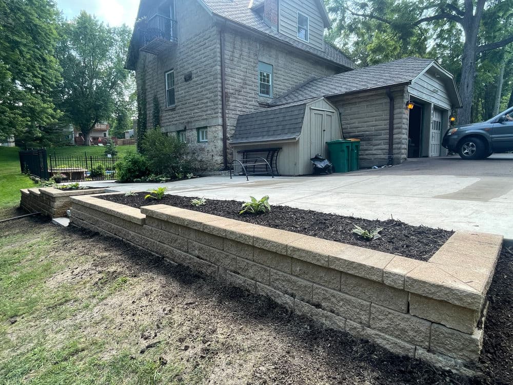 Stone retaining wall with new plants, backyard of a house, driveway, and garage visible.