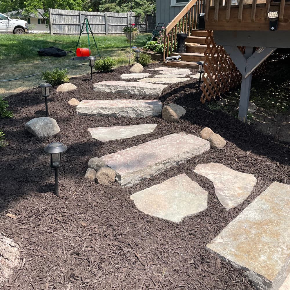 Garden pathway with stone stepping stones, mulch, and decorative lighting.