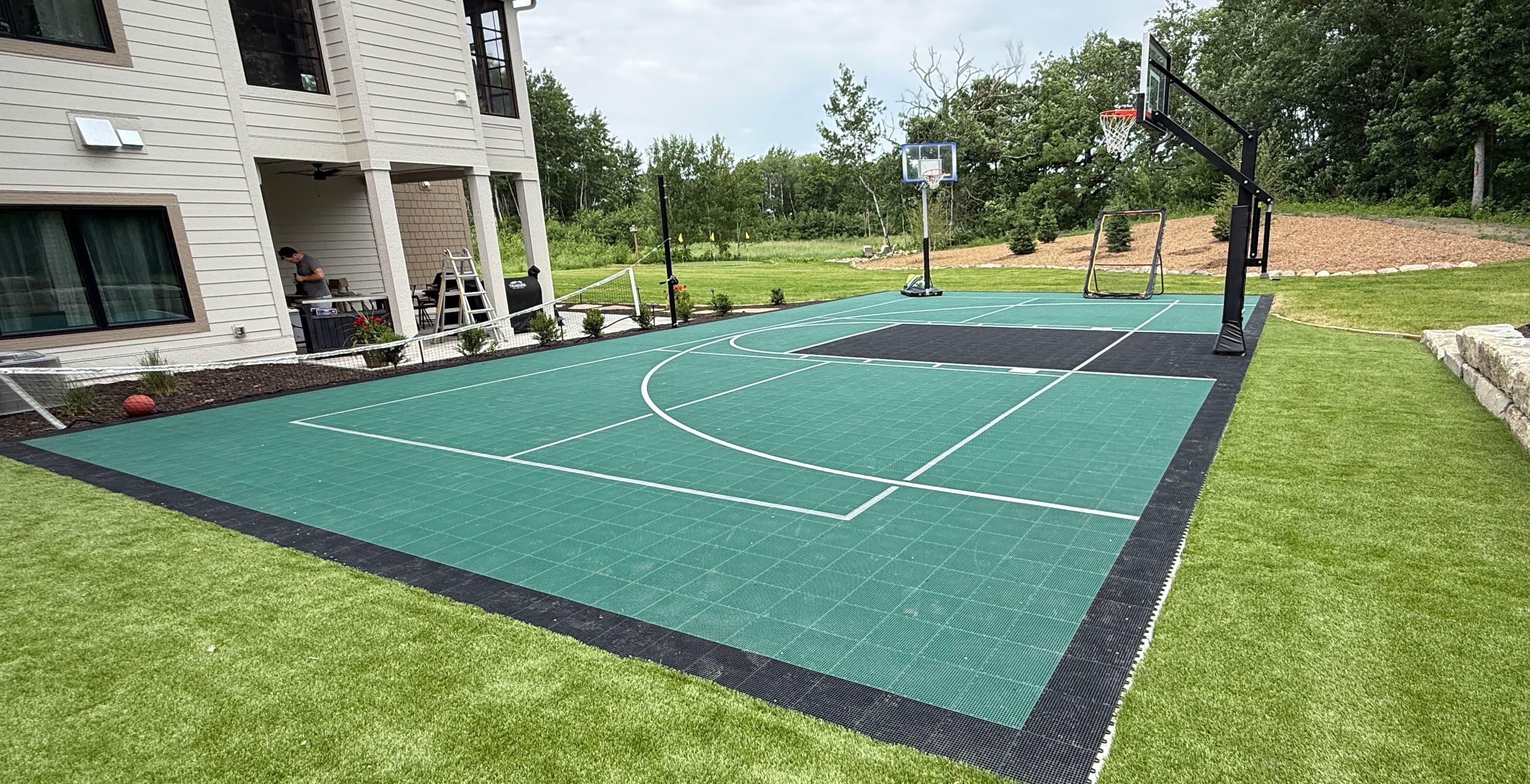Backyard basketball court with hoop, green surface, and adjacent house, surrounded by greenery.