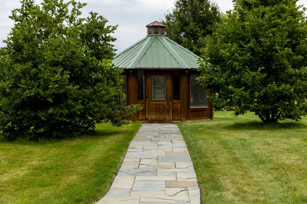 Wooden gazebo with green metal roof surrounded by trees and stone pathway.